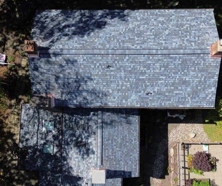 Aerial view of a house with a new shingle roof surrounded by trees and a patio area with outdoor furniture.